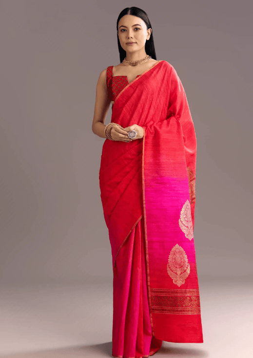 A person gracefully draped in a vibrant red and pink handloom dress featuring intricate gold patterns. The dress showcases traditional craftsmanship with detailed designs along the pallu and border, emphasizing the elegance of handloom dresses. The person accessorizes with gold jewelry, including a necklace, bangles, and a ring, complementing the rich texture of the attire. The neutral background enhances the outfit’s striking colors and weaving artistry